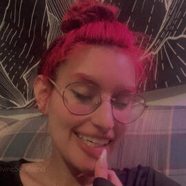 Nemea with their eyes closed smiling and their thumb with a fake pink nail pulling down their bottom lip. They have red round rimmed glasses, nose piercings and fey hair pulled up into a bun. They sit on a green couch and have a black and white tapestry b
